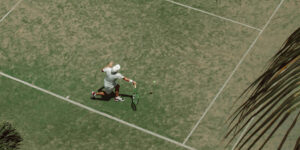 Aerial view of a man swinging a tennis racket on a grass court with palm fronds, used for a blog on sports massage for tennis and golfer’s elbow.